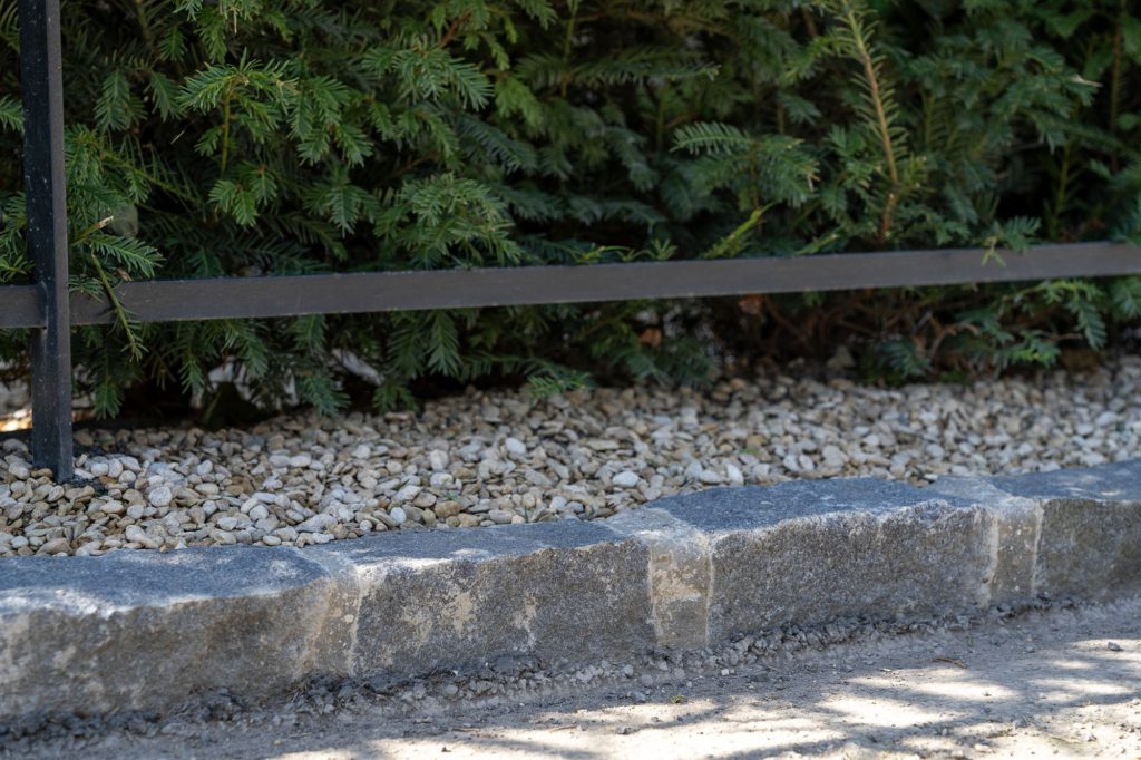 Granite setts laid as a kerb between a gravel margin and the lane, below a clipped yew hedge and black estate railing