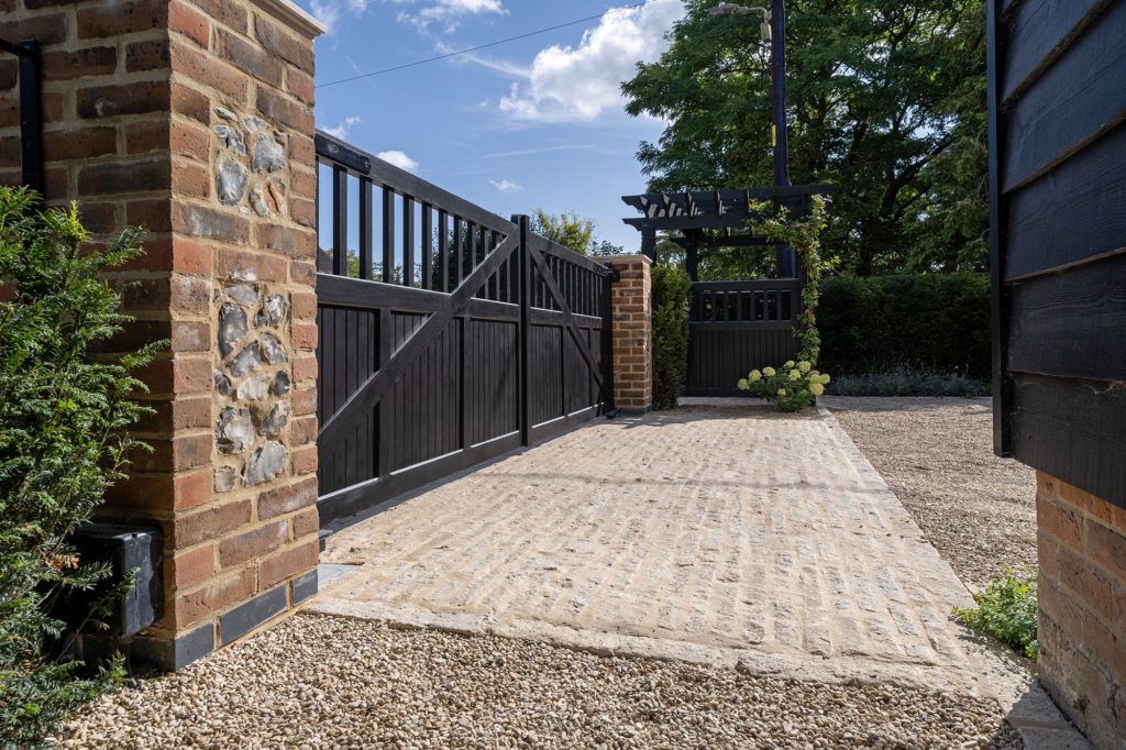 Black timber driveway gates on a brick and flint pier, with a Purbeck limestone sett path running through to the pedestrian gate