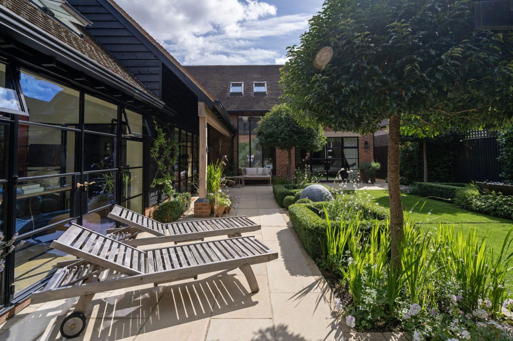 La Roche limestone paving running along a glazed barn elevation beside timber loungers, clipped box hedging and lawn
